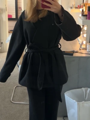 Svart omlottkappa med bälte - Säljer en svart kappa i omlottmodell med bälte i midjan. Strl xs men passar garanterat större. Från bikbok, skriv vid frågor💗