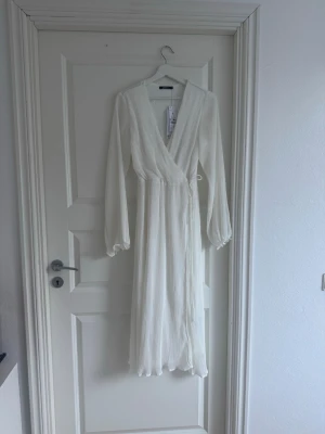 Vit omlottklänning med knytband - Lång vit omlottklänning med v-ringning och knytband i midjan. Klänningen har långärmade, lätt puffiga ärmar och är tillverkad i ett ribbat tyg som ger en snygg struktur. Perfekt för sommarens alla tillfällen.