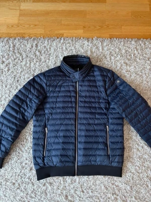 Mörkblå dunjacka från Moose Knuckles - Snygg mörkblå dunjacka från Moose Knuckles med quiltat mönster, två dragkedjeförsedda fickor och ribbade muddar vid ärmslut och nederkant. Jackan har hög krage och diskret logga på ärmen. Perfekt för kyliga dagar. Köpt på Johnells. Nypris 5699 kr. Luvan tillkommer också, glömde ha med den på bilden bara…