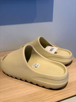 Beige Adidas Yeezy Slide tofflor - Säljer ett par beige Adidas Yeezy Slide tofflor i syntetmaterial. De har en chunky design med öppen tå och räfflad sula för extra grepp. Perfekta för chill dagar och enkel att glida i. Diskret Adidas-logga på fotbädden. Vid frågor så är det bara att höra av sig! 