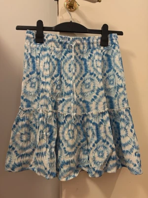 Blåvit volangkjol med batikmönster - Supersöt kort kjol i blått och vitt med batikinspirerat mönster och volang nedtill. Kjolen har resår i midjan för skön passform och är gjord i ett mjukt bomullstyg. Perfekt för sommarens alla äventyr!