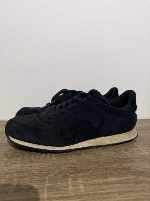 Svarta sneakers från Armani i mocka - Snygga svarta sneakers från Armani med klassisk logga på hälen. Skorna har en låg siluett, snörning och är tillverkade i mocka med vit sula och svart yttersula. Perfekta för dig som gillar stilrena och tidlösa sneakers.