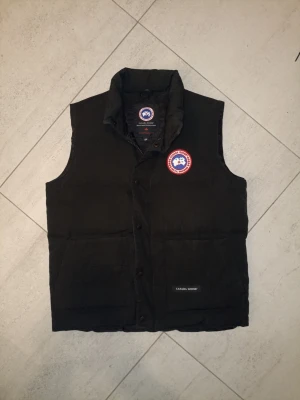 Canada Goose väst-svart - Beskrivning: Säljer en stilren och klassisk Canada Goose väst i svart. tryckknappar + dragkedja Västen är sparsamt använd och väl omhändertagen. Inga hål, fläckar eller defekter. Perfekt som lager på lager under höst/vinter eller svala vårdagar.Har använt några gånger men den är som ny. Säljer den för att jag rensar garderoben. Så hoppas någon annan har användning för den. Priset är prutbart vid snabb affär. Skickas spårbart via Plick eller möts upp enligt överens