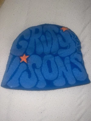 Blå mössa med tryck och stjärnor - Cool blå mössa med texten 'GROOVY VISIONS' i stora bokstäver och två orange stjärnor på framsidan. Baksidan har ett spindelnätsmönster i ljusblått. Perfekt för dig som gillar färgstarka accessoarer och unik stil.