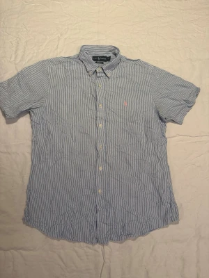 Blårandig skjorta från Ralph Lauren - Klassisk blårandig skjorta från Ralph Lauren med korta ärmar och knappstängning framtill. Skjortan har button-down krage och den ikoniska lilla logotypen broderad på bröstet. Perfekt för en avslappnad men ändå stilren look.