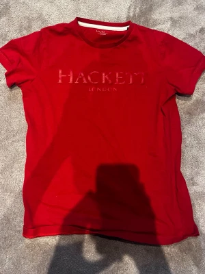 Röd Hackett London t-shirt -  300 kr Snygg röd t-shirt från Hackett London med diskret logga framtill. Klassisk passform och rund halsringning. Perfekt till jeans eller shorts för en clean och enkel stil.