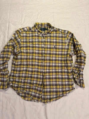 Rutig skjorta från Ralph Lauren XXL - Säljer en klassisk rutig skjorta från Ralph Lauren i färgerna gul, vit, svart och röd. Skjortan har button-down krage, långa ärmar och broderad logga på bröstet. Tillverkad i mjuk bomull och har en avslappnad classic fit.