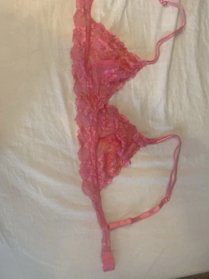 Rosa spets-bh med blommönster - Säljer en söt rosa bh i triangelformad modell med tunna axelband och hakknäppning bak. Bh:n är gjord i genomskinlig spets med blommönster och har dekorativa kanter. Perfekt för dig som gillar romantiska detaljer och lätt material. Aldrig använd men har tagit bort prislappen