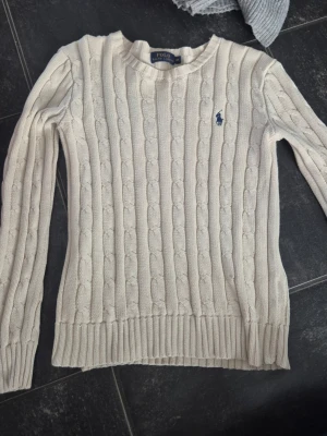 Vit kabelstickad tröja Polo Ralph Lauren - Kabelstickad vit tröja från Polo Ralph Lauren med rund halsringning och blå broderad logga på bröstet. Långa ärmar och ribbade muddar vid ärmslut och nederkant. Klassisk och tidlös design i mjukt material.