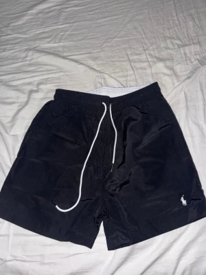 Svarta badbyxor från Polo Ralph Lauren - Snygga svarta badbyxor från Polo Ralph Lauren med vit dragsko i midjan och klassisk logga broderad på vänster ben. De har elastisk midja och innerbyxa i mesh för extra komfort. Perfekta för stranden eller poolen. Storlek S. Aldrig använda. Kan ge billigare på snabb affär
