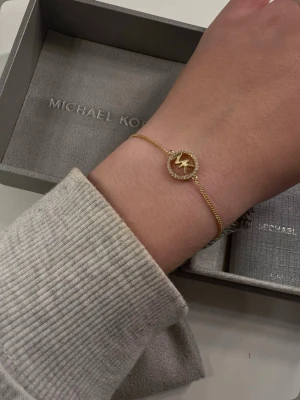 Guldigt armband Michael Kors - Snyggt guldigt armband från Michael Kors med en rund berlock. Tunn kedja och justerbar längd med små detaljerade brickor i ändarna. Ny utan prislapp