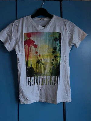 Vit California t-shirt från H&M - Säljer en vit t-shirt från H&M med färgglatt tryck av palmer och texten 'California' på framsidan. T-shirten har korta ärmar.  Perfekt för dig som gillar avslappnad och cool stil.