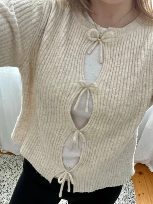 Beige cardigan med knyt - Trendigt, beige, ribbstickad cardigan med fyra dekorativa knyt framtill och öppna detaljer. Långärmad och croppad modell. Perfekt att styla över ett linne eller t-shirt. 