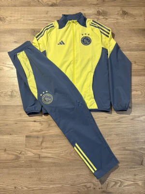 Adidas Ajax Tracksuit - Ajax träningsset från Adidas i gul och blå färg. Jackan har dragkedja, ståkrage och klubbmärke på bröstet. Byxorna har resår i midjan och klubbmärke på låret. Setet är gjort i lätt och funktionellt material, perfekt för träning. Storlek S passar även M