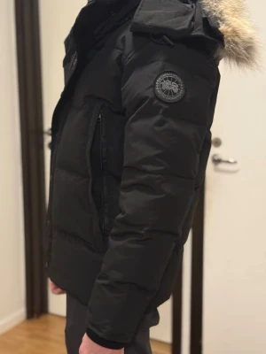 Svart dunjacka från Canada Goose - Säljer en svart dunjacka från Canada Goose med huva och avtagbar fuskpäls. Jackan har patch-logga på ärmen, dragkedja och knappar framtill samt två stora fickor. Perfekt för kalla vinterdagar och riktigt snygg streetstil.