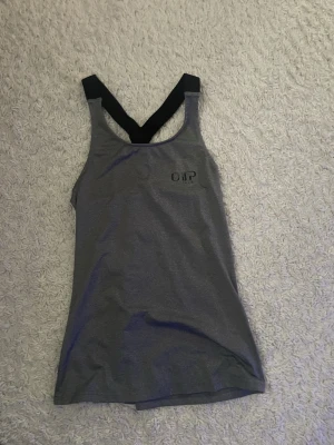 Grå linnetopp med svarta band - Snygg grå linnetopp med svarta, breda axelband i racerback-modell. Toppen är ärmlös och har en sportig look, perfekt för dig som gillar enkel och stilren design. Materialet känns mjukt och följsamt mot kroppen.