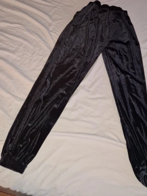 Svarta mjukisbyxor i sammet - Säljer ett par svarta mjukisbyxor i mjuk sammet. Byxorna har resår i midjan och vid bensluten för en skön och avslappnad passform. Perfekta för chill dagar eller när du vill vara extra bekväm.