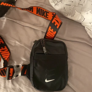 Svart axelväska från Nike - Snygg svart axelväska från Nike med justerbar axelrem i svart och orange med stora NIKE-loggor. Väskan har ett vitt Nike-märke framtill, dragkedja upptill och ett extra fack på framsidan. Perfekt för att bära det viktigaste när du är på språng.