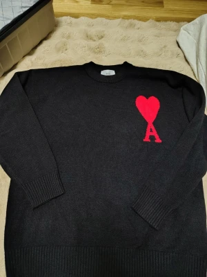 Svart stickad tröja Ami Paris hjärta - Svart stickad tröja från Ami Paris med röd hjärtdetalj och bokstaven A på bröstet. Tröjan har rund halsringning, långa ärmar och ribbstickade muddar vid ärmslut och nederkant. Perfekt för dig som gillar stilrena plagg med en twist.