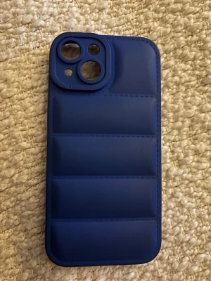 Blått vadderat mobilskal till iPhone 15 - Snyggt blått mobilskal med vadderad design och räfflade sektioner. Skalet har stora utskärningar för kamerorna och är gjort i ett mjukt, flexibelt material som ger extra grepp och skydd. Perfekt för dig som vill ha en stilren och modern look på din iPhone.