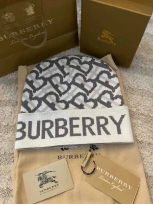 Vit och grå Burberry mössa - Snygg vit och grå mössa från Burberry med ikoniskt monogrammönster och stor logotyp framtill. Mössan har uppvikt kant och är stickad i mjukt material, perfekt för att hålla värmen och ge en exklusiv touch till din outfit.