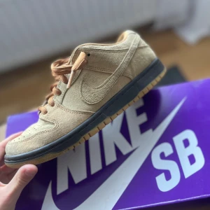 Nike SB Dunk Low i beige mocka - Nike SB Dunk Low sneakers i beige mocka med svarta och gummifärgade sulor. Skorna har klassisk snörning och finns med både ljusbruna och mörkbruna skosnören. Perfekta för dig som gillar streetwear och skate-vibe.
