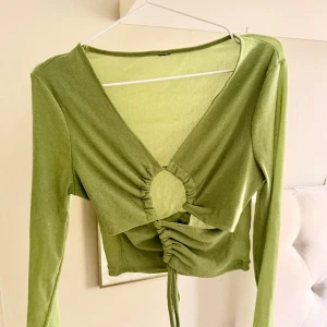 Grön glittrig croppad topp med snörning - Snygg ljusgrön långärmad topp med glittrigt tyg och croppad passform. Toppen har djup v-ringning, en rund cut-out framtill och snörning som ger en cool rynkad effekt. Perfekt för dig som vill sticka ut med en trendig och unik look. Fel storlek för mig 