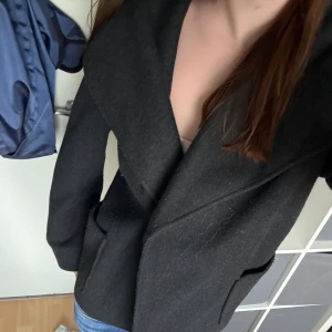 Svart omlott kappa med bred krage - Snygg svart kappa med bred krage och omlott-design. Jackan har två stora fickor framtill och en stilren, lite loose passform. Perfekt för dig som gillar minimalistisk stil och vill ha något som funkar till jeans eller dressat. Materialet känns som ull och ger en mjuk känsla. Perfekt till hösten/ tidig vinter och knäpps med knapp. Fanns snöre men borttappat 💘💘Storlek M men passar även mig med S/Xs. Nypris 749.
