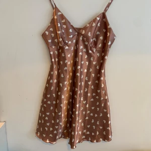 Ljus brun hjärtklänning med smala axelband - Supersöt kort ljus brun klänning med vita hjärtan och smala axelband. Klänningen har en lätt glansig finish och v-ringad urringning framtill. Perfekt för dig som gillar lekfulla mönster och en feminin siluett.