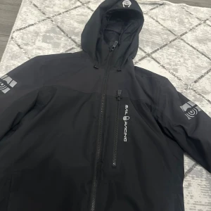 Svart pufferjacka från Sail Racing - Säljer en svart pufferjacka från Sail Racing med huva och dragkedja. Jackan har logga på bröstet och ärmen, samt quiltad insida för extra värme. Perfekt för kyliga dagar och har en clean, sportig look.