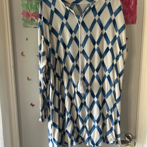 Blåvit rutig långärmad klänning - Superfin långärmad klänning från Miss Alice med blått och vitt rutmönster. Klänningen har snörning med tofs framtill och är i en loose passform. Perfekt för dig som gillar unika mönster och avslappnad stil.