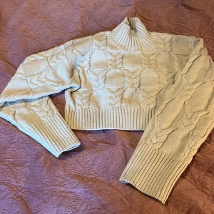 Beige stickad polotröja från Monki - Säljer en beige stickad polotröja från Monki i storlek S. Tröjan har ett snyggt flätmönster, ribbade muddar och hög krage. Perfekt för lager-på-lager och ger en mysig vibe till din outfit.
