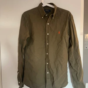 skjorta från Polo Ralph Lauren - Olivgrön skjorta från Polo Ralph Lauren med klassisk button-down krage och vit knappstängning. Liten orange logobroderi på bröstet. Skjortan är långärmad och tillverkad i mjuk bomull, perfekt för en chill och stilren look.