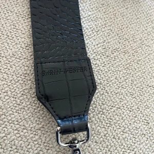 Svart axelrem från Carin Wester - Snygg och bred svart axelrem från Carin Wester med croco-präglad yta och blanka metalldetaljer. Perfekt att använda till din favoritväska för att ge den en cool och modern vibe. Remmen har karbinhakar i silver för enkel fastsättning.
