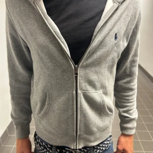 Grå hoodie från Polo Ralph Lauren - Grå hoodie från Polo Ralph Lauren med dragkedja framtill, två fickor och broderad logga på bröstet. Mjuk insida och klassisk huva. Perfekt för chill dagar och enkel att matcha med jeans. Den har en snygg design. Storleken är ungefär xs-xxs. Priset kan diskuteras👍