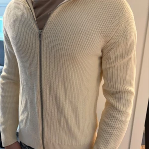 Vit stickad kofta från East West - Säljer en stilren vit stickad kofta från East West i storlek M. Koftan har ribbad struktur, hel dragkedja framtill och långärmad passform. Perfekt för lager-på-lager och enkel att matcha med olika outfits.