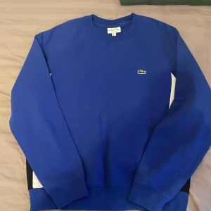 Blå sweatshirt från Lacoste - Blå sweatshirt från Lacoste med relaxed fit och klassisk krokodillogga på bröstet. Tröjan har långa ärmar, rund hals och vita samt svarta paneler på sidorna. Perfekt för dig som gillar stilren design och sportig vibe.