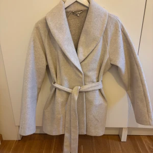 Beige kappa med bälte från ONLY - Snygg beige kappa från ONLY i storlek S. Jackan har en bred krage, knytskärp i midjan och är tillverkad i ett mjukt material som känns lyxigt. Perfekt för dig som gillar stilrena och enkla plagg med en modern touch.