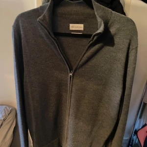 Grå merinoullskofta från These Glory Days - Snygg grå kofta i merinoull från These Glory Days. Den har hel dragkedja framtill, hög krage och lång ärm. Perfekt för lager-på-lager och enkel att matcha med det mesta. Stilren och tidlös design som funkar året runt.