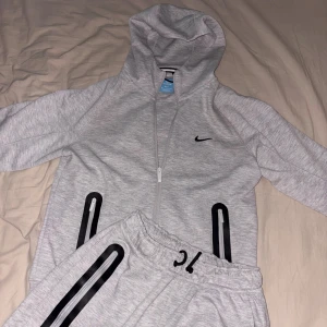 Nocta tech fleece grå - Snygg ljusgrå hoodie från Nike med hel dragkedja och svart logga på bröstet. Tröjan har huva, långa ärmar och svarta detaljer vid fickorna. Perfekt för träning eller chill. Materialet är mjuk bomull och polyesterblandning.