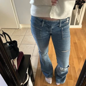 pleated flare jeans  - Säljer ett par ljusblå bootcut jeans med snygga slitningar på knäna och råa kanter. Jeansen har låg midja, dubbla knappar i linningen och stora framfickor. Perfekta för dig som gillar en avslappnad och trendig stil. Ginas trendiga jeans 