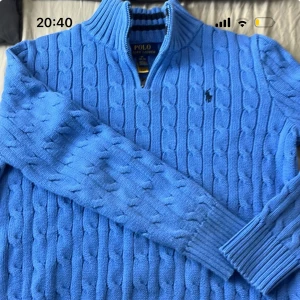 Blå stickad tröja Polo Ralph Lauren - Säljer en blå kabelstickad tröja från Polo Ralph Lauren med halv dragkedja och hög krage. Tröjan har lång ärm, ribbade muddar och den klassiska broderade loggan på bröstet. Perfekt för dig som gillar stilren och tidlös design.