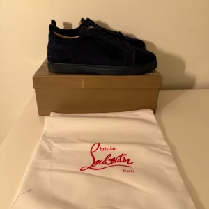 Mörkblå mocka sneakers från Louboutin - Snygga mörkblå sneakers från Christian Louboutin i lyxig mocka med klassisk röd sula och svarta detaljer. Skorna har rund tå, snörning och diskret broderad logga på hälen. Perfekta för dig som vill sticka ut med exklusiv stil.