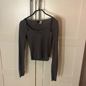Grå ribbad långärmad topp från H&M - Säljer en grå ribbad långärmad topp från H&M Divided. Toppen har en croppad passform, spetskant vid halsringningen och en liten rosett framtill. Materialet är mjukt och stretchigt, perfekt för en chill och snygg look.