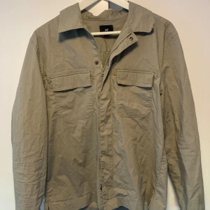 Beige overshirt fmed dragkedja - Snygg beige overshirt från H&M i bomull med klassisk krage, två bröstfickor med lock och både dragkedja och tryckknappar framtill. Perfekt lager-på-lager-plagg med lång ärm och avslappnad passform. Perfekt jacka nu till hösten, passar dig som är runt 175cm. Aldrig använd.