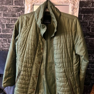 Olivgrön quiltad jacka från Truenorth - Snygg olivgrön quiltad jacka från Truenorth med dragkedja och tryckknappar framtill. Jackan har ståkrage, långa ärmar och två fickor med dragkedja. Perfekt för kyliga dagar och har en stilren, sportig look.