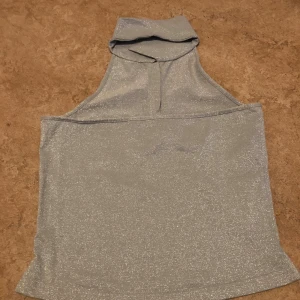 Glittrig silver polotopp - Snygg ärmlös polotopp i glittrigt silvertyg. Toppen har hög krage och rak passform, perfekt för att sticka ut. Materialet är stretchigt och skimrar i ljuset. Enkel men ändå riktigt trendig design.