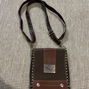 Brun axelväska från Giorgio Armani - Säljer en brun axelväska från Giorgio Armani i skinn med croco-mönstrad yta och silverfärgade nitar längs kanterna. Väskan har justerbar axelrem, lock med metallogo och dragkedja inuti. Perfekt för dig som vill ha en unik och stilren accessoar.