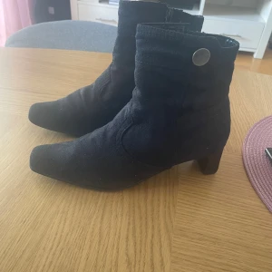 Svarta mocka stövletter med klack Park West stl 37 - Snygga svarta stövletter i mocka med markerad klack. Skorna har dragkedja på insidan och en dekorativ knappdetalj upptill. Perfekta för dig som vill ha en stilren och trendig look till höst och vinter.