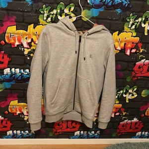 Grå hoodie från Burberry med rutig huva - Snygg grå hoodie från Burberry i bomull med dragkedja och klassiskt rutigt mönster i huvan. Tröjan har två fickor framtill, snörning i huvan och ribbade muddar. Perfekt för dig som vill ha en stilren och lyxig vardagslook. Passar perfekt till dig som 185 till 180 cm lång. 